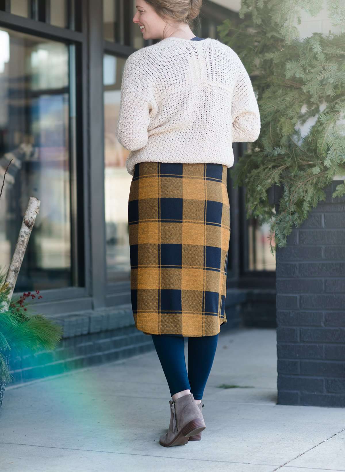 Woman wearing a modest plaid sweater style midi skirt. This skirt has an elastic waist and a tie front. It is paired with tights and short ankle boots.