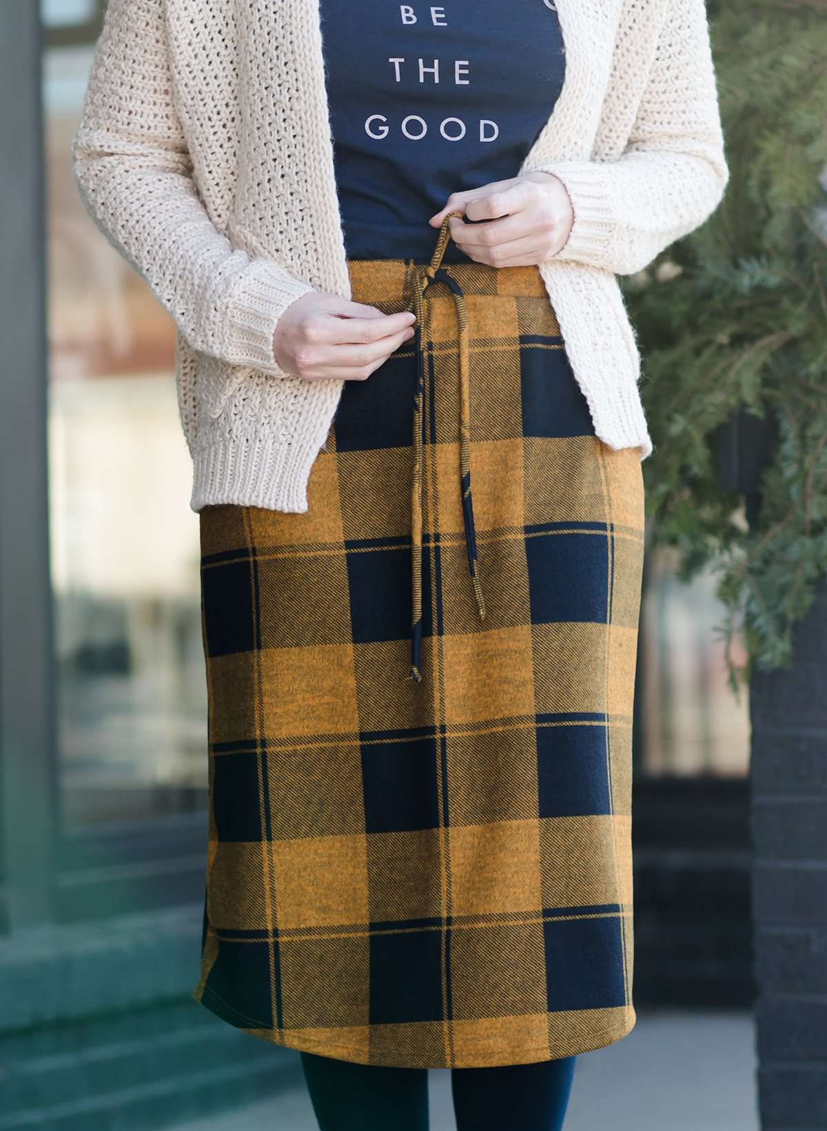 Woman wearing a modest plaid sweater style midi skirt. This skirt has an elastic waist and a tie front. It is paired with tights and short ankle boots.