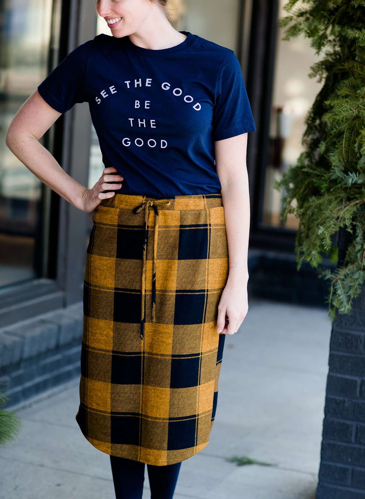 Woman wearing a modest plaid sweater style midi skirt. This skirt has an elastic waist and a tie front. It is paired with tights and short ankle boots.