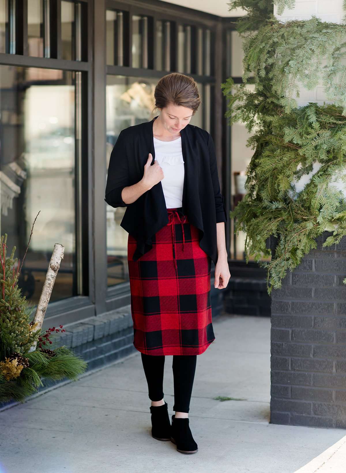 Woman wearing a modest plaid sweater style midi skirt. This skirt has an elastic waist and a tie front. It is paired with tights and short ankle boots.