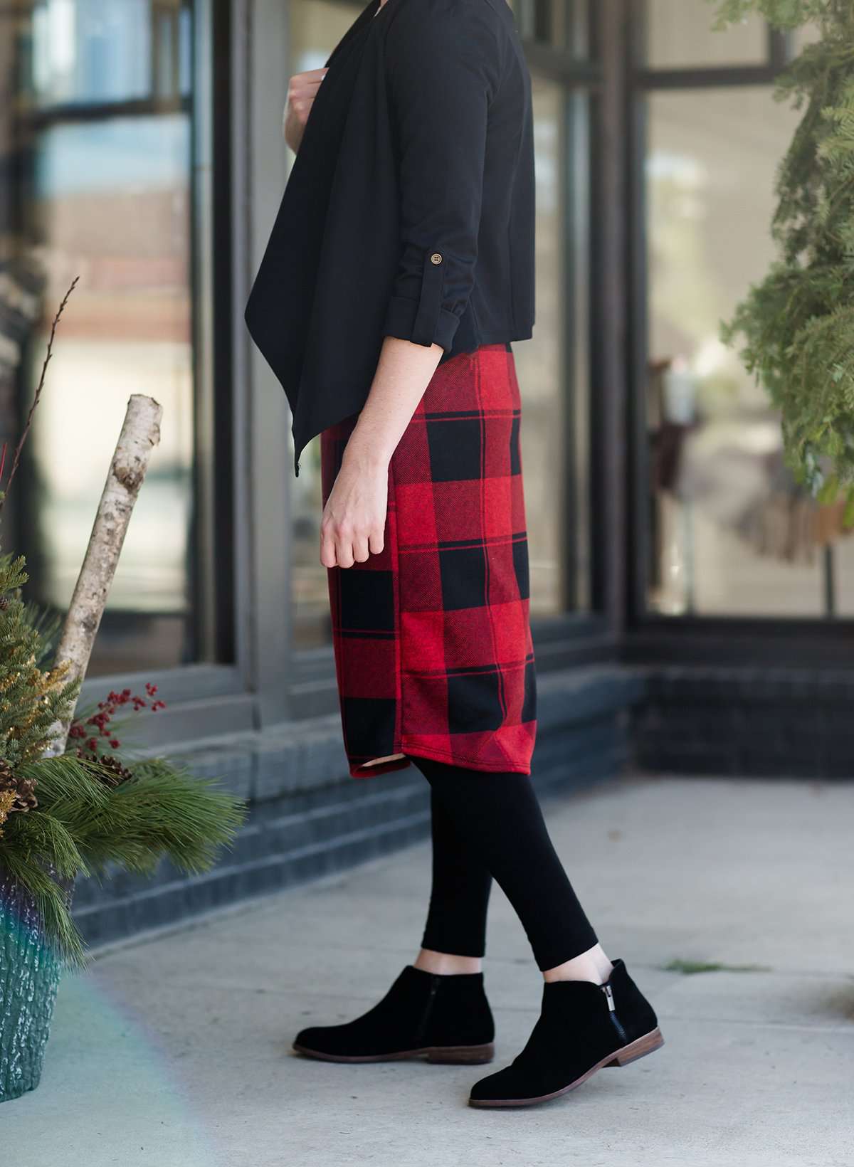 Woman wearing a modest plaid sweater style midi skirt. This skirt has an elastic waist and a tie front. It is paired with tights and short ankle boots.