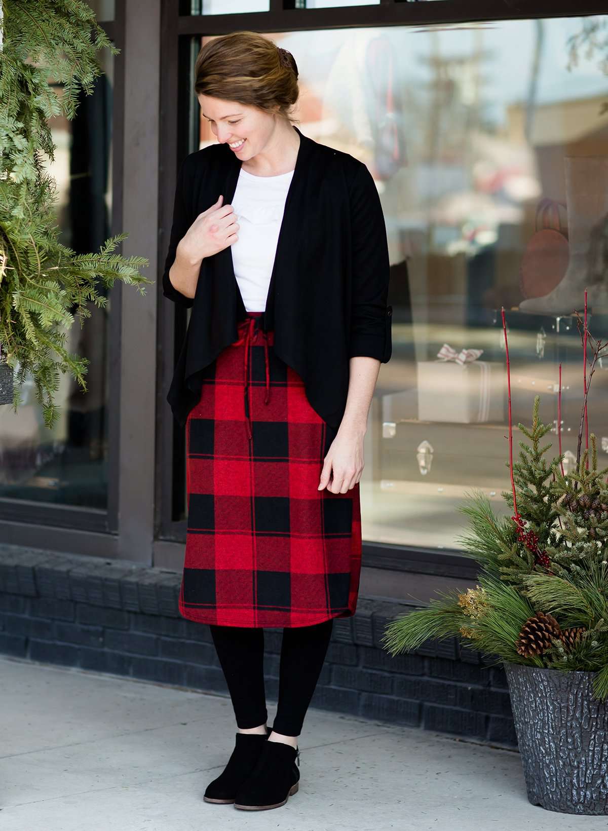 Woman wearing a modest plaid sweater style midi skirt. This skirt has an elastic waist and a tie front. It is paired with tights and short ankle boots.