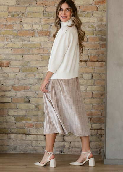 Tan pink velvet pleated midi skirt shown at an angle, styled with an ivory funnel neck sweater and heels.