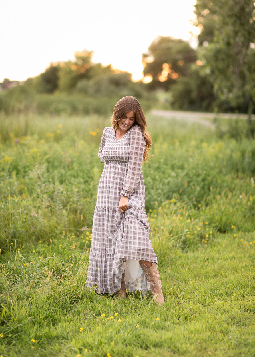 A woman stands in a grassy field at sunset wearing a long-sleeve, tiered maxi dress with a gray and white windowpane plaid pattern.