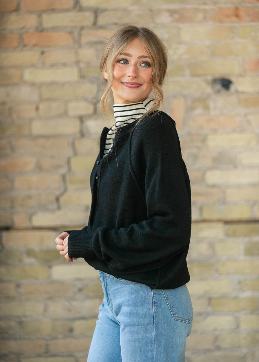 Black henley sweater layered over a striped turtleneck, styled with wide-leg denim and black flats.