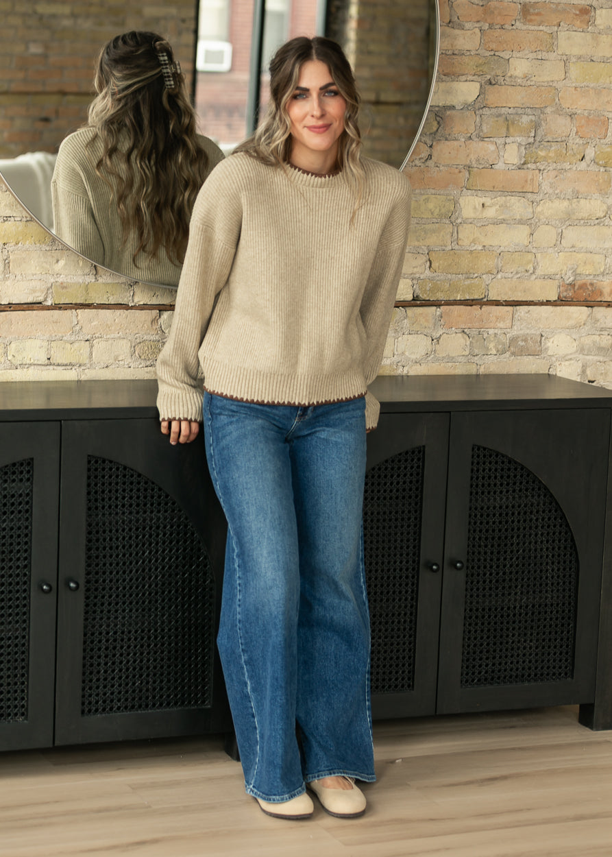 A woman stands indoors wearing a beige knit sweater and blue wide-leg jeans, leaning against a black cabinet in front of a brick wall and mirror.