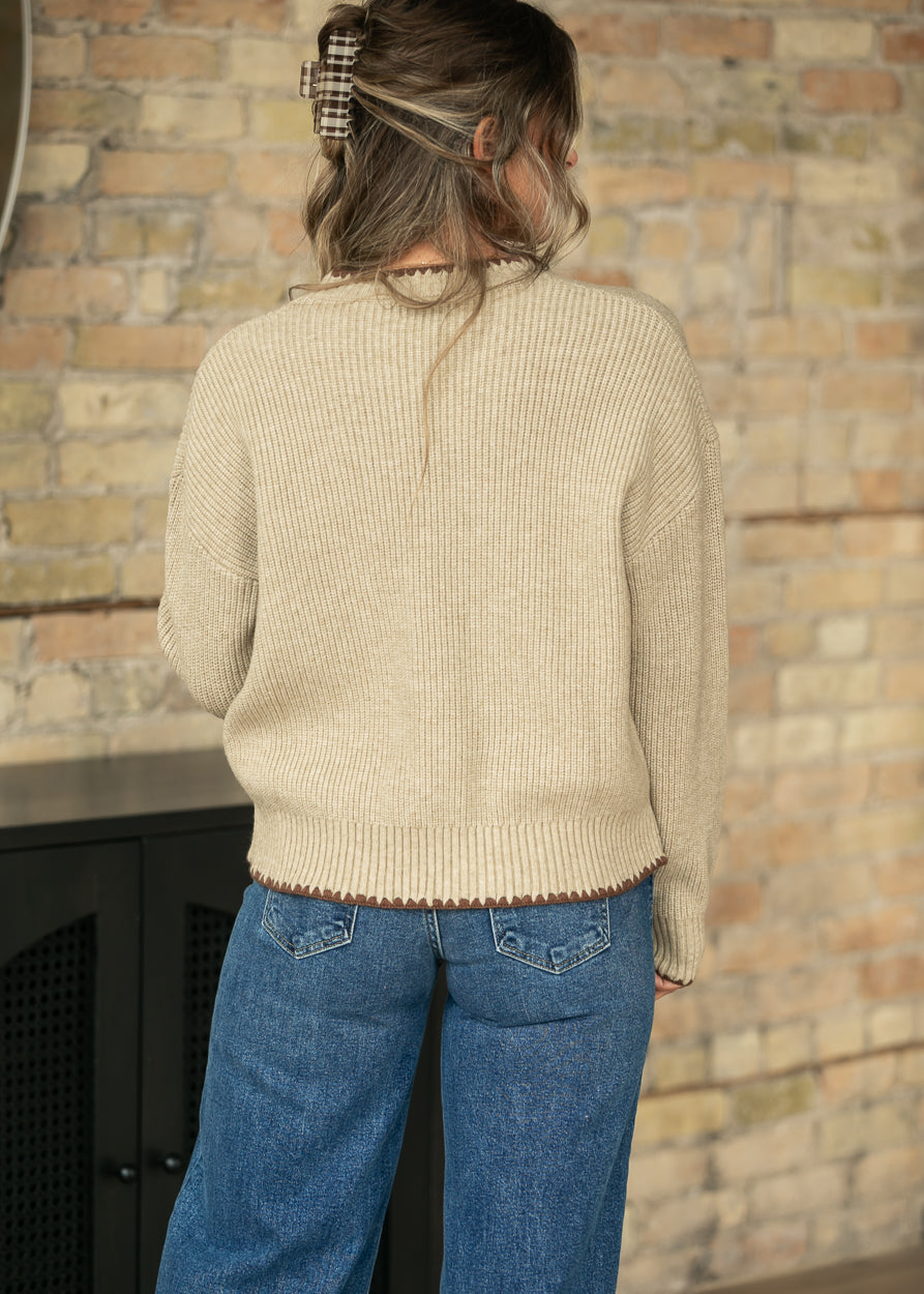 A woman viewed from behind wearing a cream-colored ribbed knit sweater and blue jeans, standing in front of a brick wall.