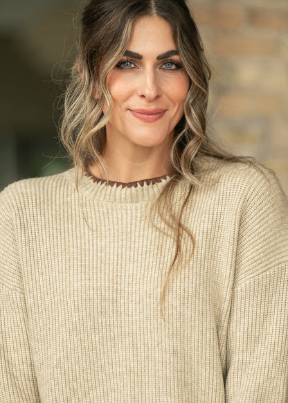 A woman with wavy blonde-highlighted hair wearing a beige ribbed knit sweater with a dark brown scalloped neckline, photographed against a softly blurred outdoor background.
