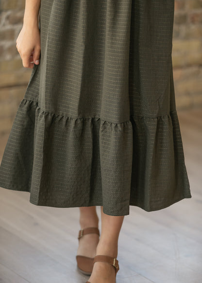 An olive green tiered midi skirt with a subtle textured pattern, shown on a model wearing brown strappy sandals against a rustic brick wall and light wood floor background.