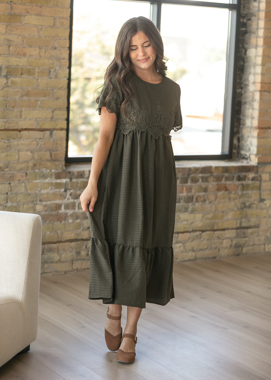 A woman wearing a dark olive green midi dress with a lace bodice and tiered skirt, standing in a room with a brick wall and large window.