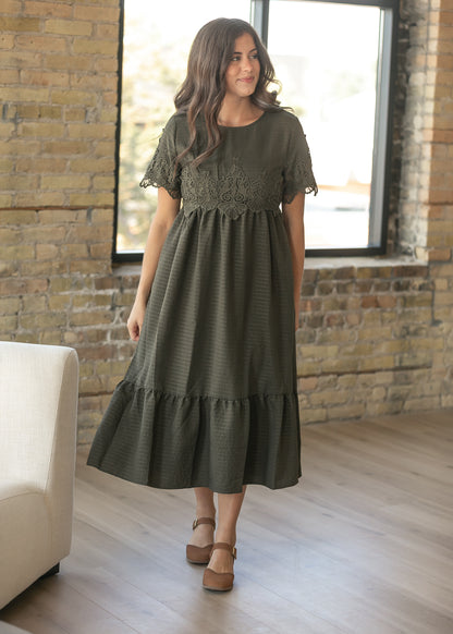 A woman wearing an olive green midi dress with a lace bodice and tiered skirt, standing in a room with a brick wall and large window.