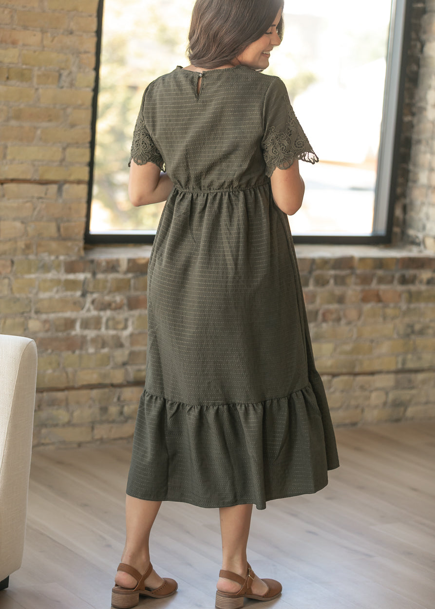 A woman wearing an olive green tiered midi dress with lace short sleeves, shown from the back in a room with a brick wall and large window.
