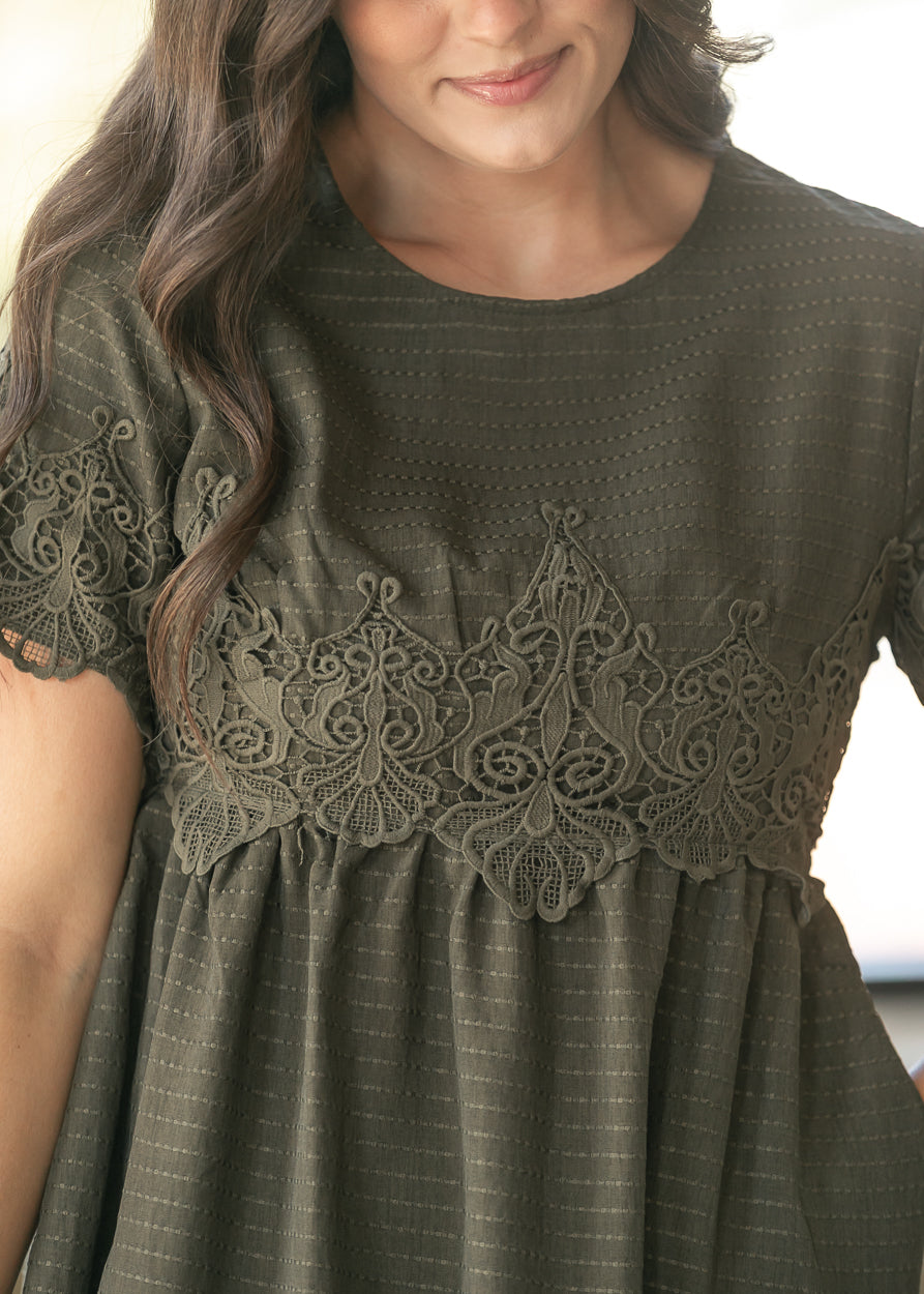 A close-up of a woman wearing an olive green short-sleeve dress with a textured striped fabric and a wide matching lace yoke across the chest.