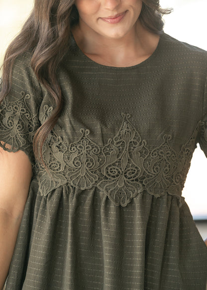 A close-up of a woman wearing an olive green short-sleeve dress with a textured striped fabric and a wide matching lace yoke across the chest.