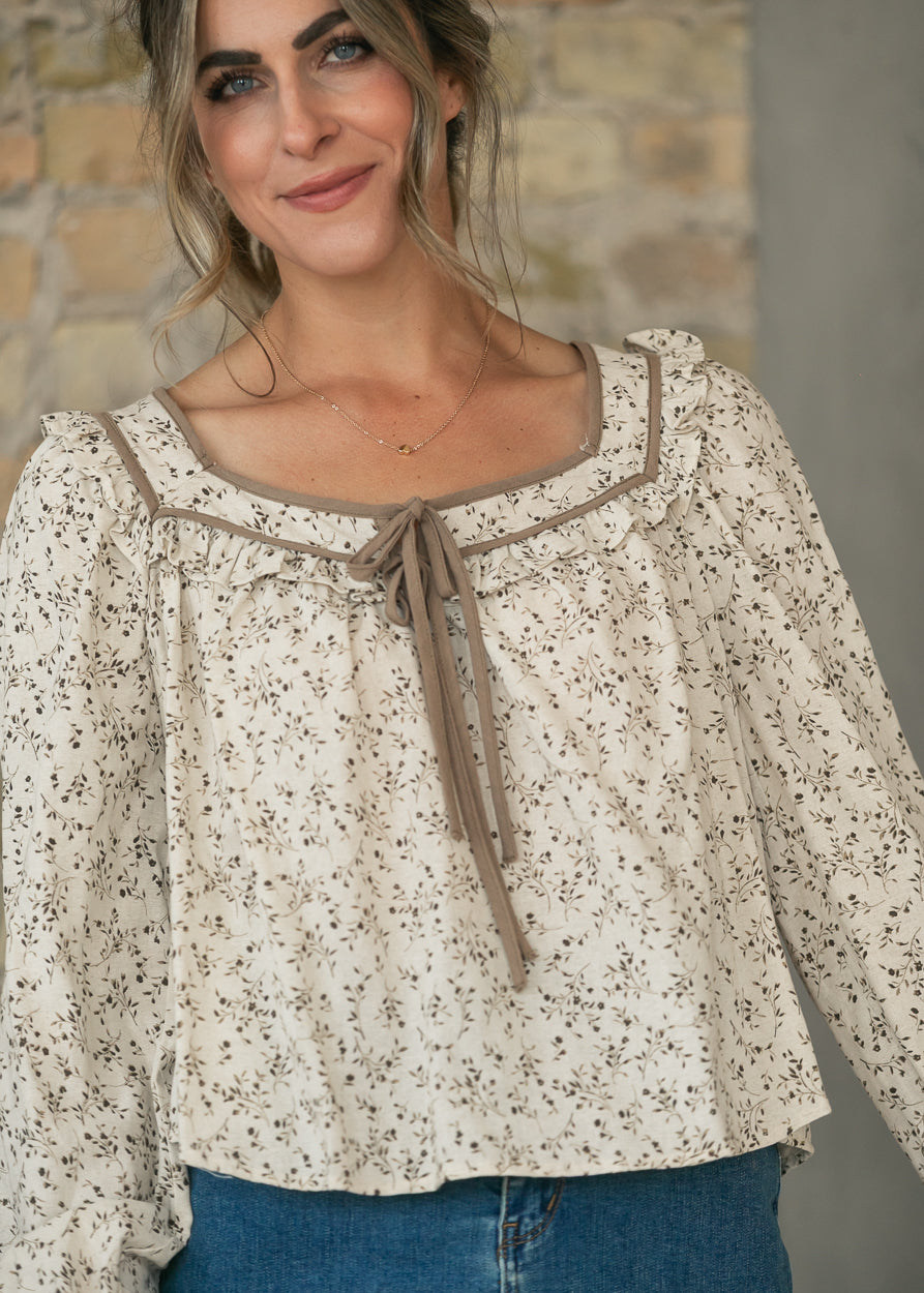 Cream botanical blouse with brown contrast trim, ruffled square neckline, and front tie detail.
