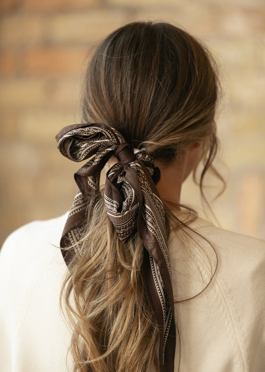 Brown bandana-print triangle scarf tied in a bow around a low ponytail.