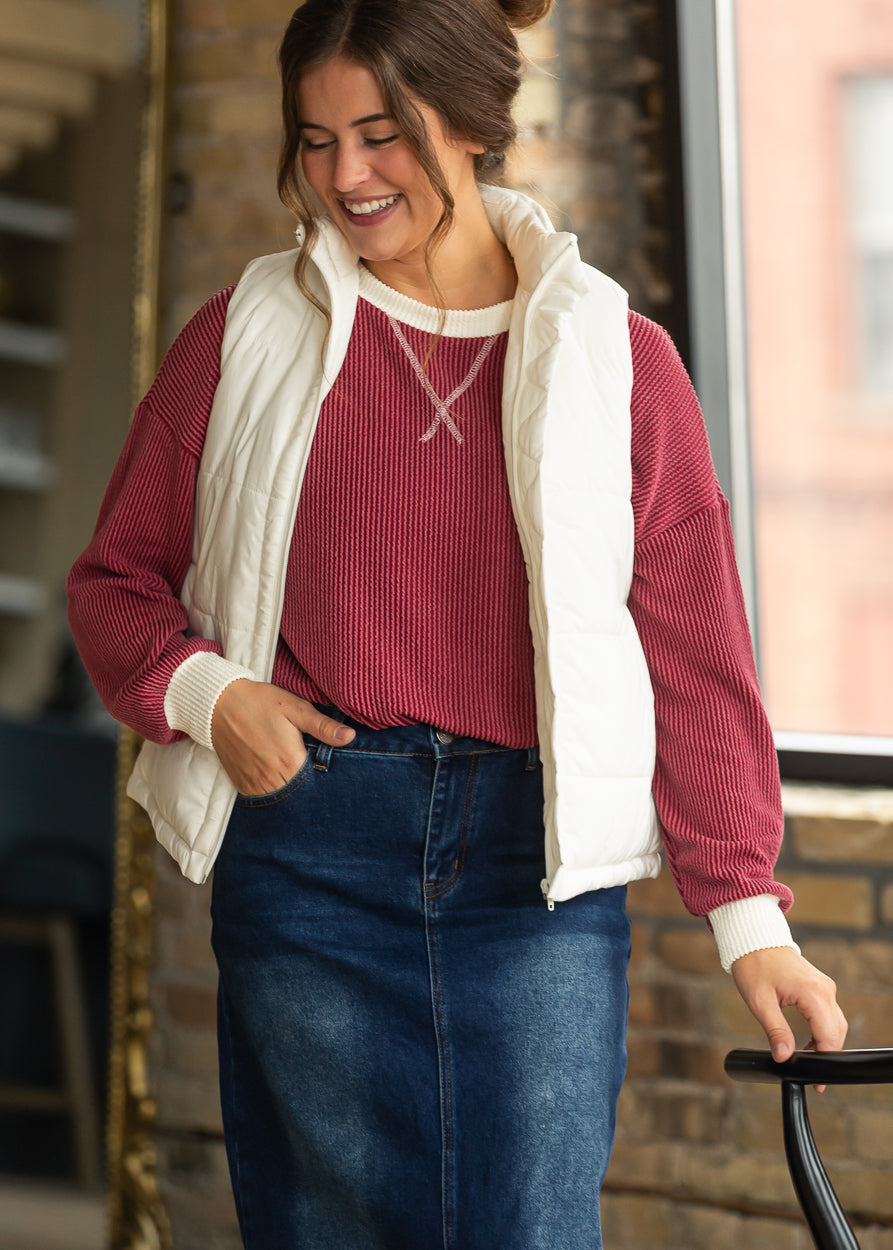 Burgundy ribbed pullover layered under a cream puffer vest with a dark wash denim maxi skirt.