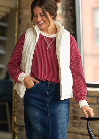 Burgundy ribbed pullover layered under a cream puffer vest with a dark wash denim maxi skirt.