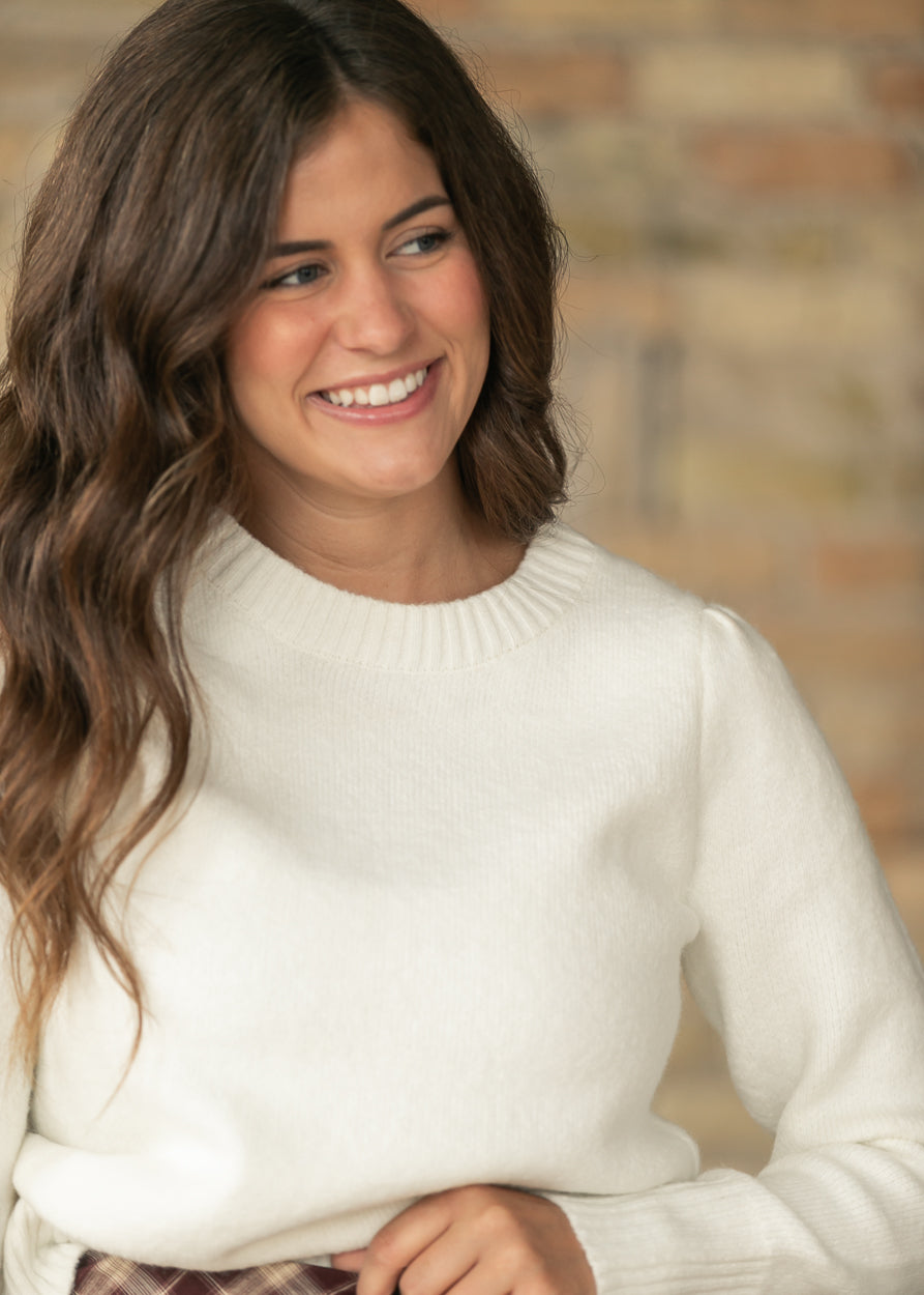 A smiling woman with long wavy brown hair wearing a cream-colored knit crewneck sweater, set against a softly blurred brick wall background.