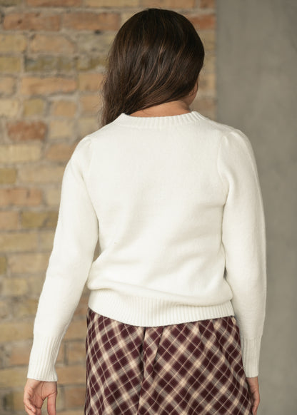 A woman viewed from behind wearing a cream-colored knit sweater and a brown and beige plaid skirt, standing in front of a rustic brick wall.