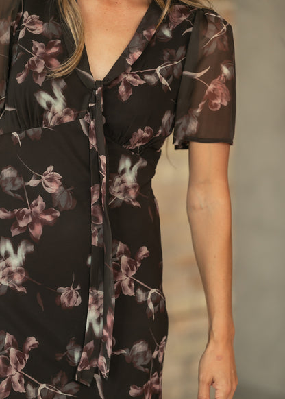 Close-up of charcoal floral maxi dress showing V-neckline, front tie, gathered waist, and sheer short sleeve.