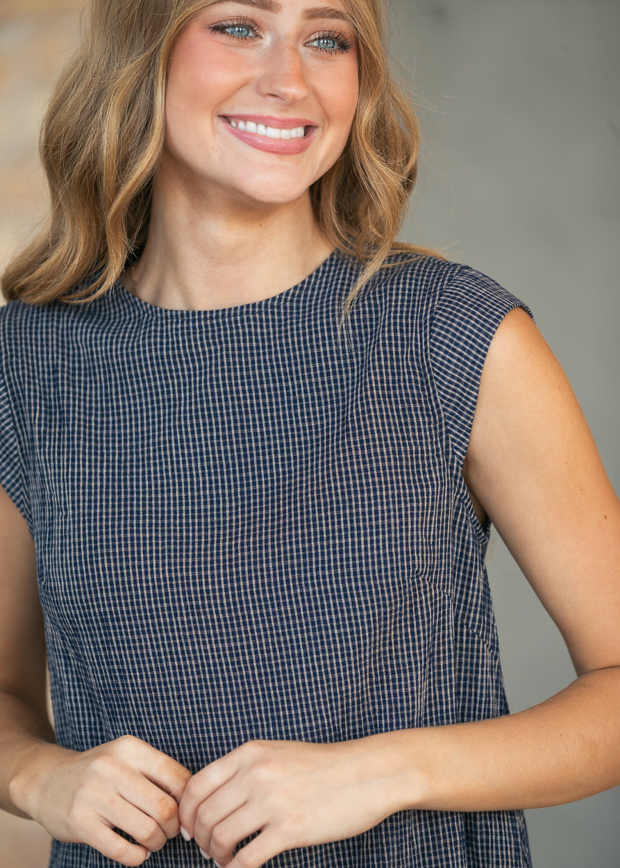 A woman wearing a navy and white micro-checkered sleeveless top, smiling and posing against a neutral gray background.