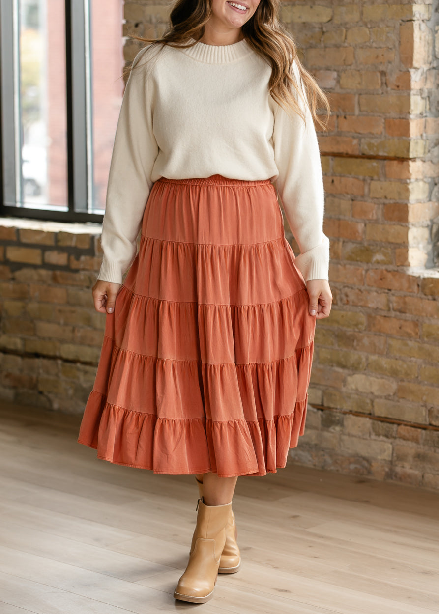 Washed rust tiered midi skirt with elastic waist and flowing silhouette, styled with an ivory sweater.