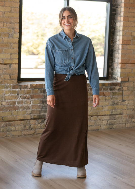 Chocolate brown knit maxi skirt with a no-slit hem, styled with a denim button-up and neutral ankle boots.
