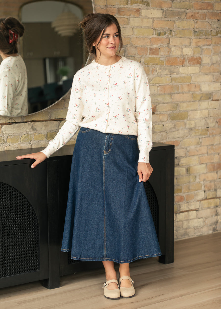 Dark indigo A-line denim midi skirt styled with a cream floral cardigan and neutral Mary Jane flats.