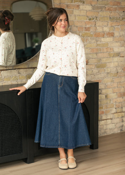Dark indigo A-line denim midi skirt styled with a cream floral cardigan and neutral Mary Jane flats.