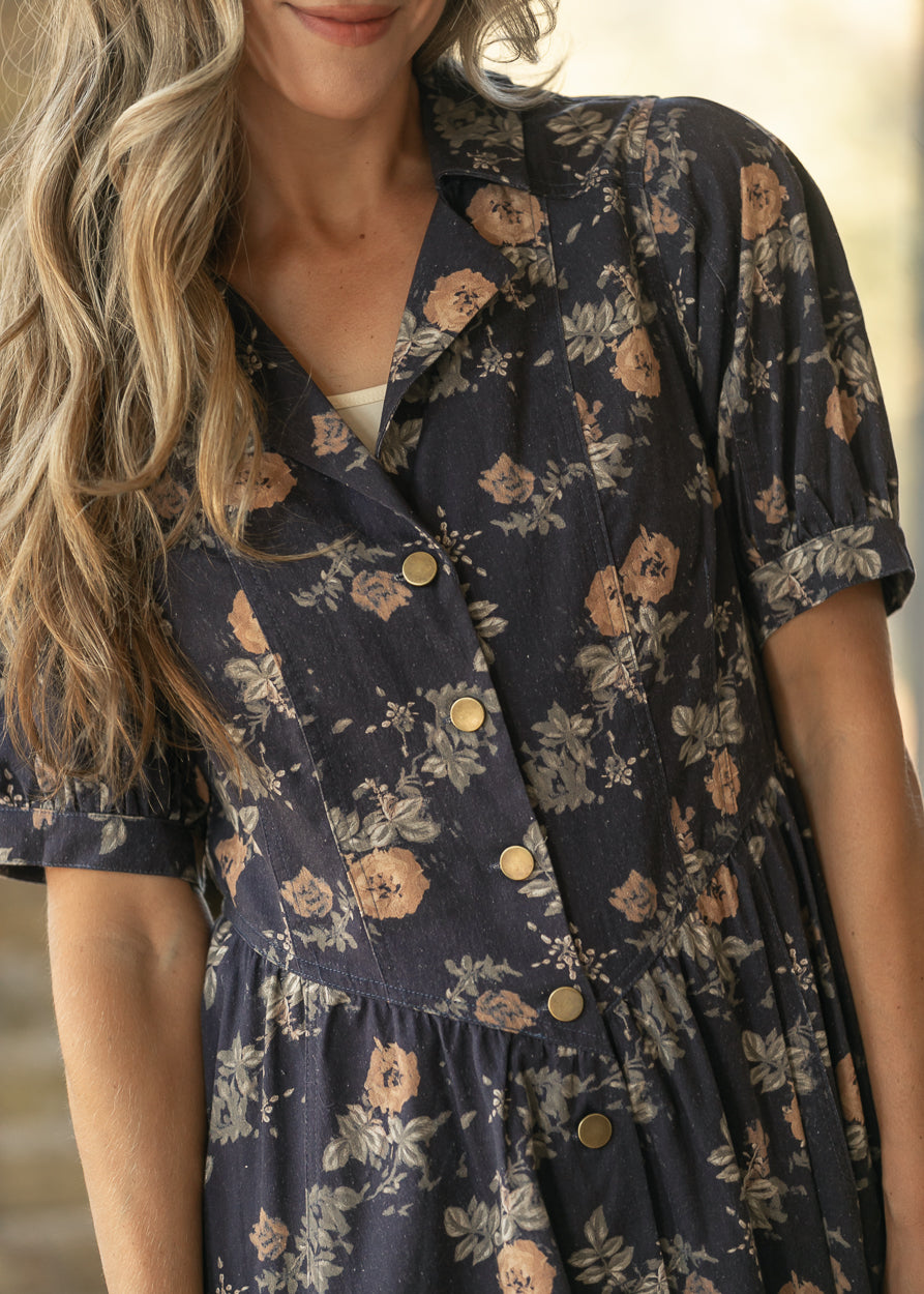 A close-up of a woman wearing a navy blue short-sleeve button-front dress with a beige and brown floral print.