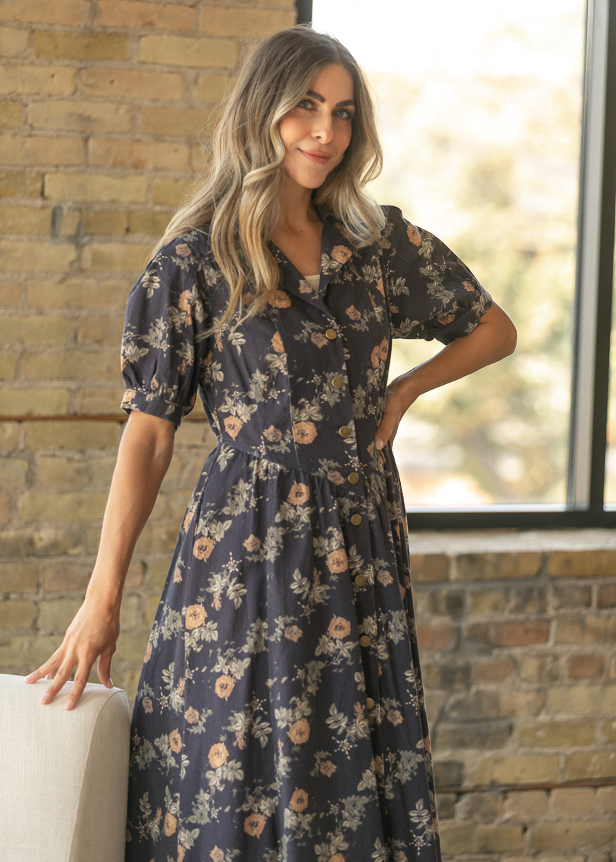 A woman wearing a dark navy floral midi dress with puff sleeves, standing in front of a brick wall window.