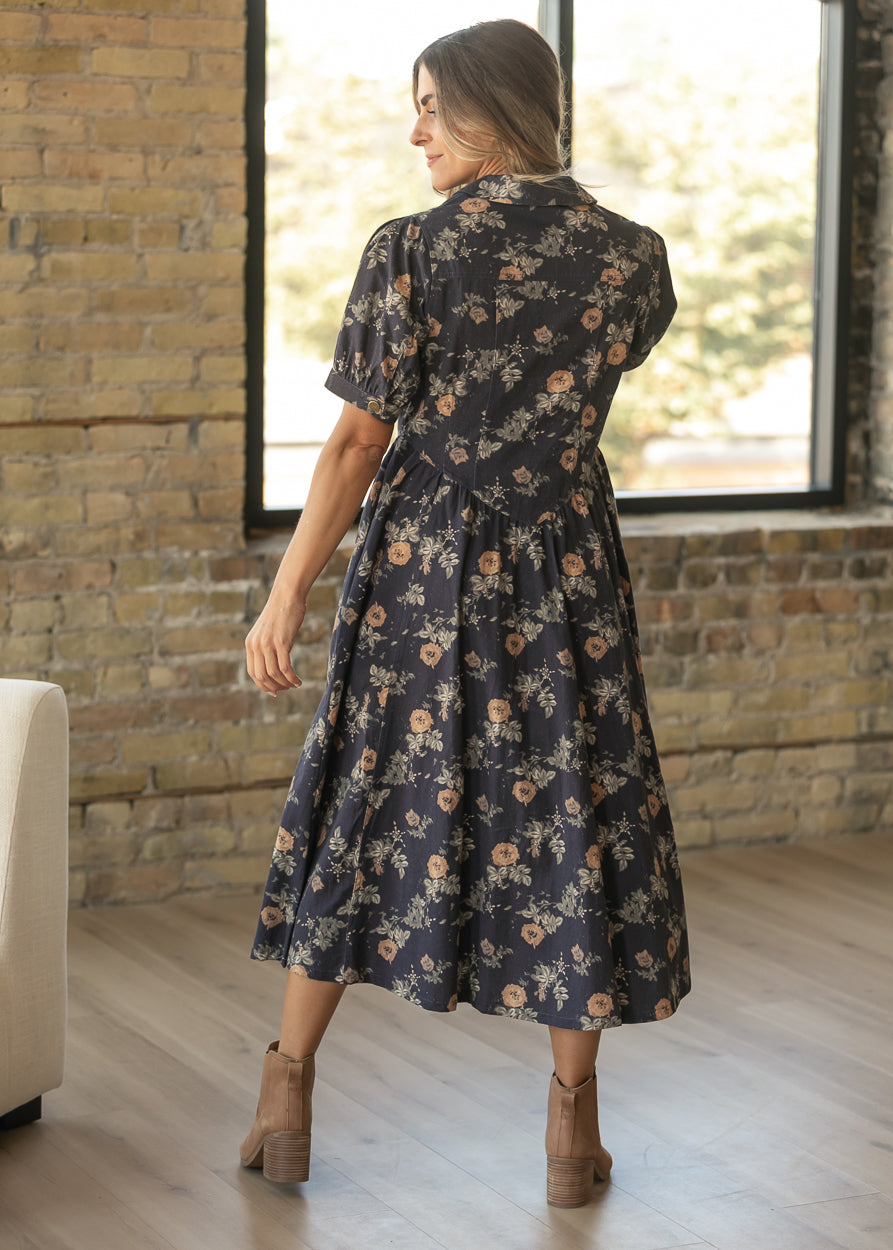 A woman wearing a dark navy midi dress with a beige and orange floral pattern, shown from the back in a loft-style room with brick walls and a large window.
