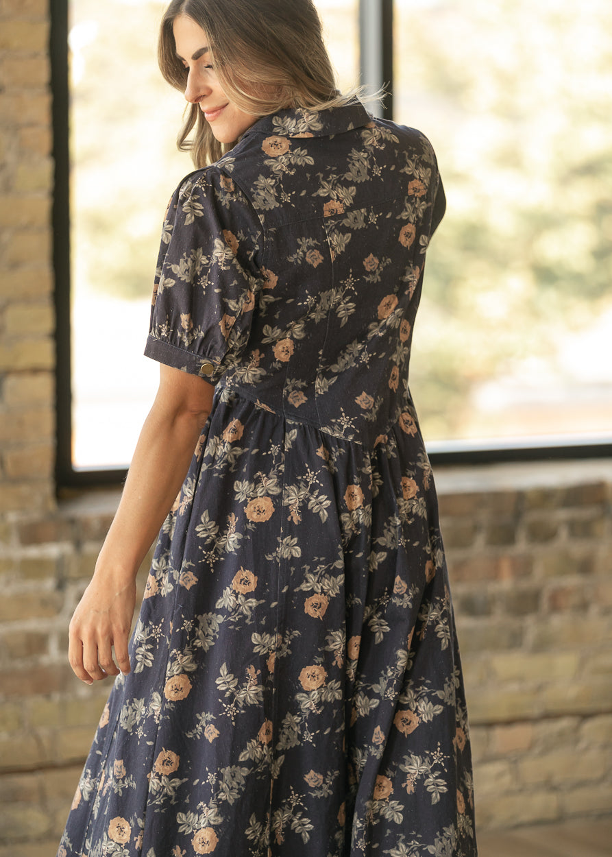 A woman wearing a dark navy floral midi dress with puff sleeves, shown from the back in a bright room with a brick wall and window.
