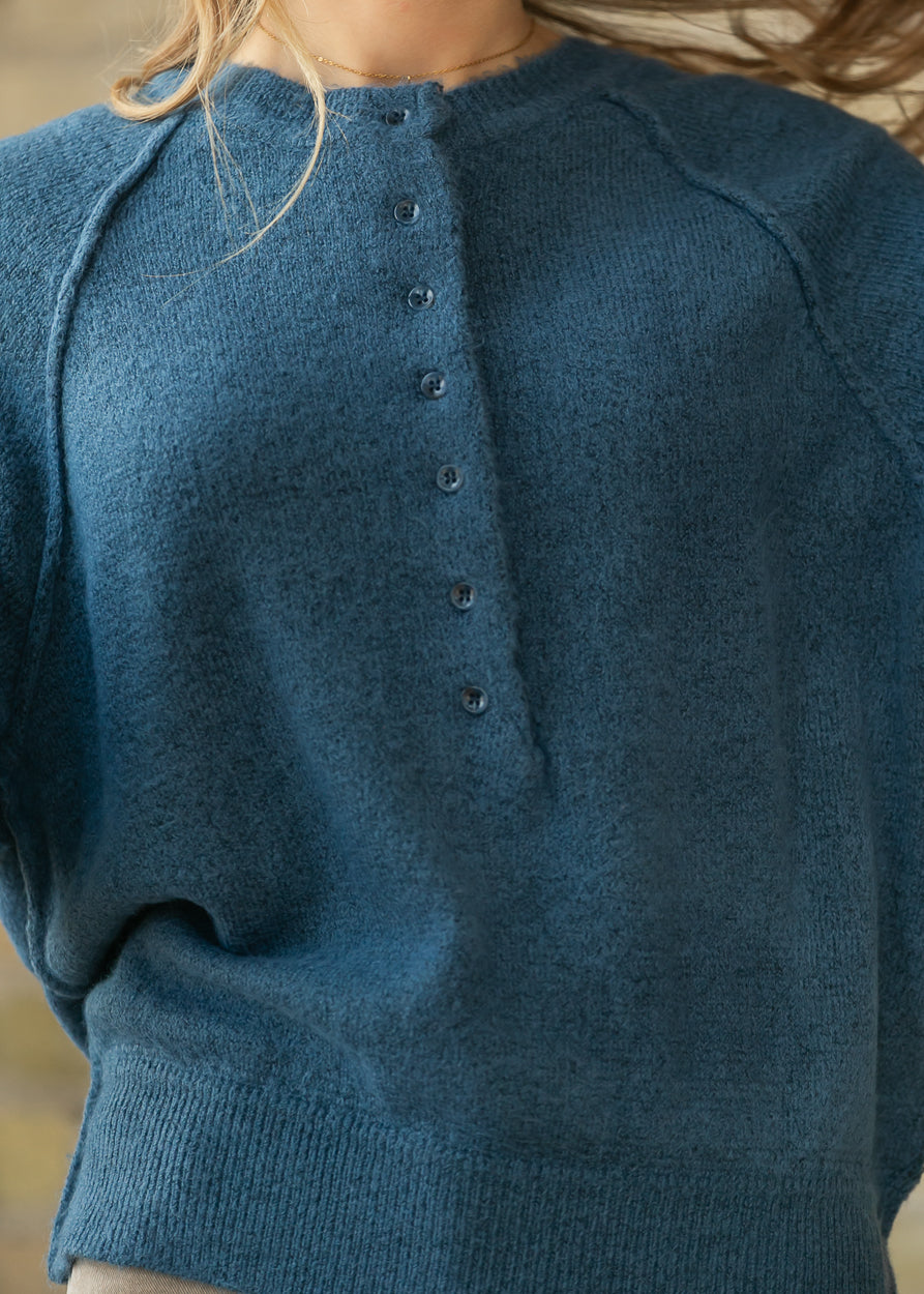 Close-up of the dusty blue henley sweater's button placket and raw-edge collar.