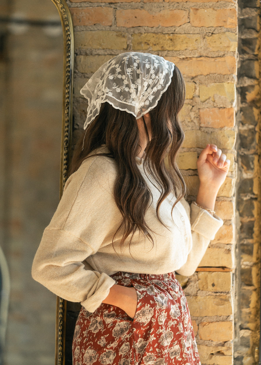 Ivory lace triangle scarf worn over dark hair, styled with a cream cardigan and floral skirt.