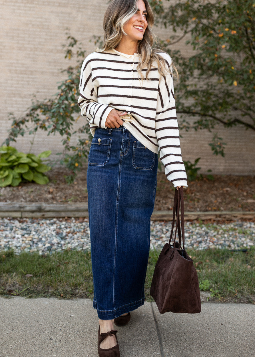 A woman wearing a cream and black striped long-sleeve top and a dark blue denim maxi skirt, holding a brown suede tote bag outdoors on a sidewalk.
