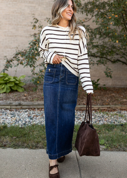 A woman wearing a cream and black striped long-sleeve top and a dark blue denim maxi skirt, holding a brown suede tote bag outdoors on a sidewalk.