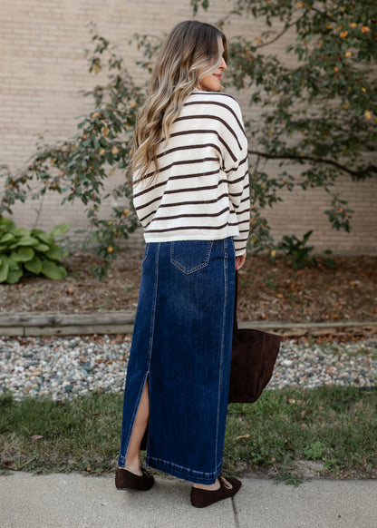 A woman stands outdoors in profile wearing a cream and brown horizontally striped knit sweater and a long dark blue denim skirt with a front slit. She is holding a dark brown tote bag and wearing brown flat shoes, set against a background of a brick wall, greenery, and gravel.