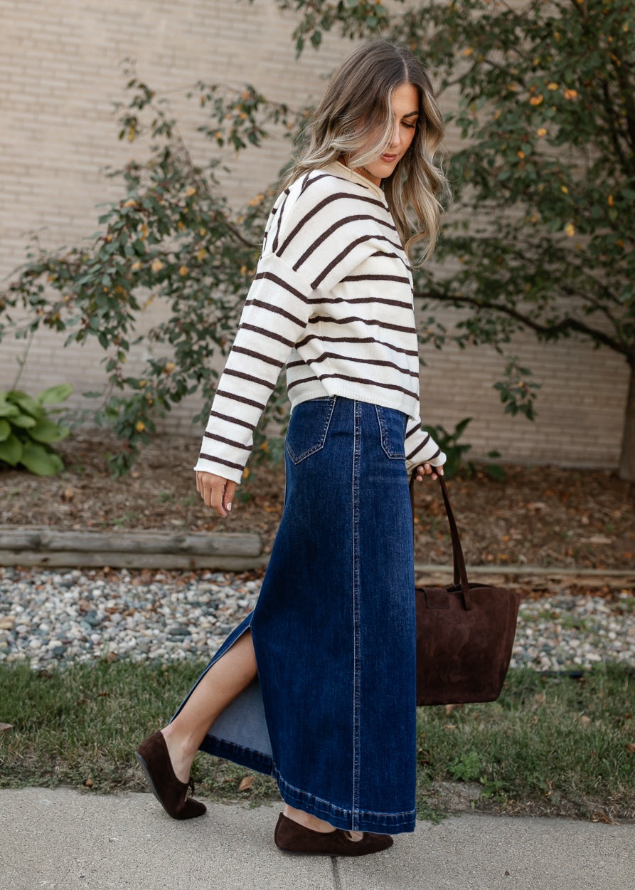 A woman wearing a cream and brown striped knit long-sleeve top and a high-waisted blue denim maxi skirt with a side slit, walking outdoors while carrying a brown leather tote bag.