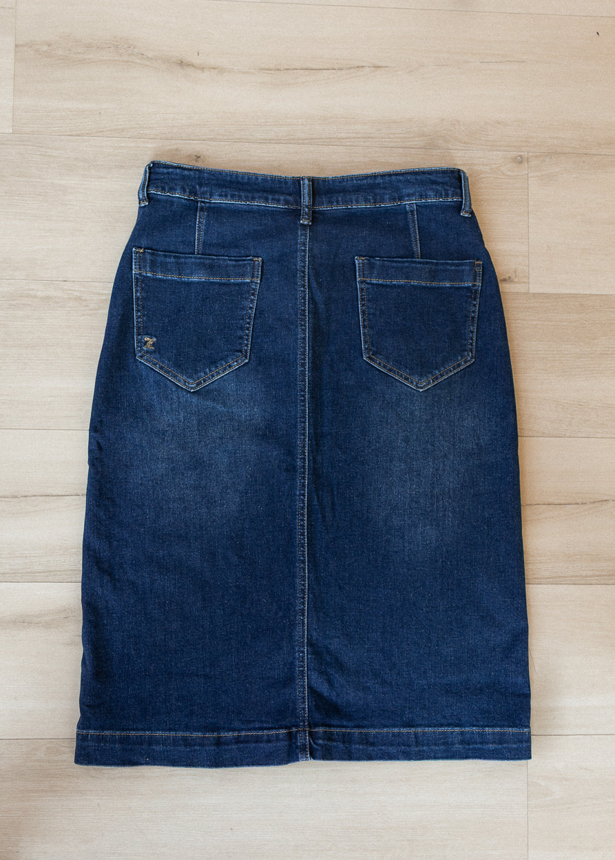 A dark blue denim skirt with two back patch pockets, laid flat on a light wood plank floor.