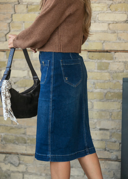 A person wearing a medium-wash blue denim pencil skirt with back pockets, paired with a brown knit sweater and holding a black shoulder bag, standing against a light brick wall.