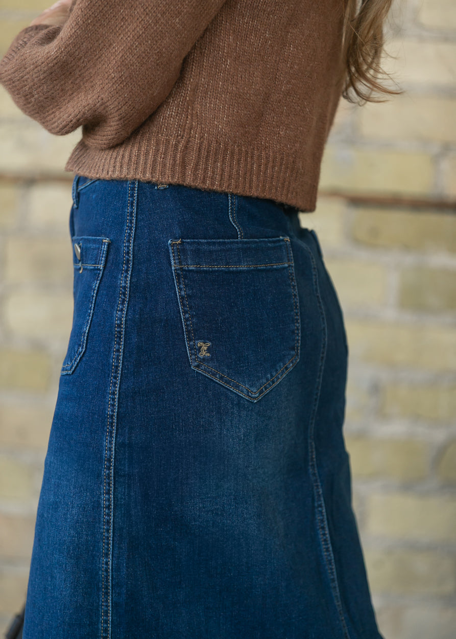 A close-up side view of a person wearing a brown ribbed knit sweater and a dark blue denim skirt, with a light brick wall in the background.