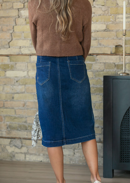 A woman shown from behind wearing a brown knit sweater and a dark blue denim midi skirt with back patch pockets, standing in front of a brick wall.