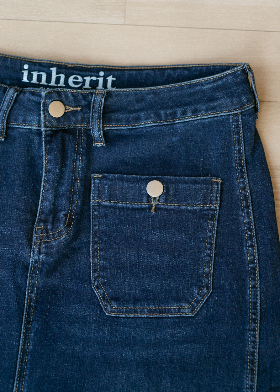Close-up of dark blue denim jeans laid flat on a light wood floor, showing the waistband with the word 'inherit' in white text and a front patch pocket with a metal button.