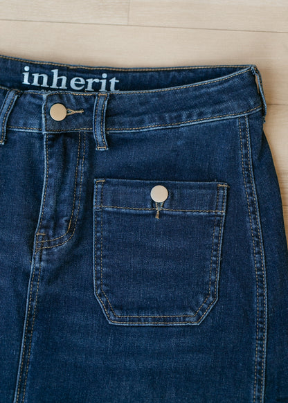 Close-up of dark blue denim jeans laid flat on a light wood floor, showing the waistband with the word 'inherit' in white text and a front patch pocket with a metal button.