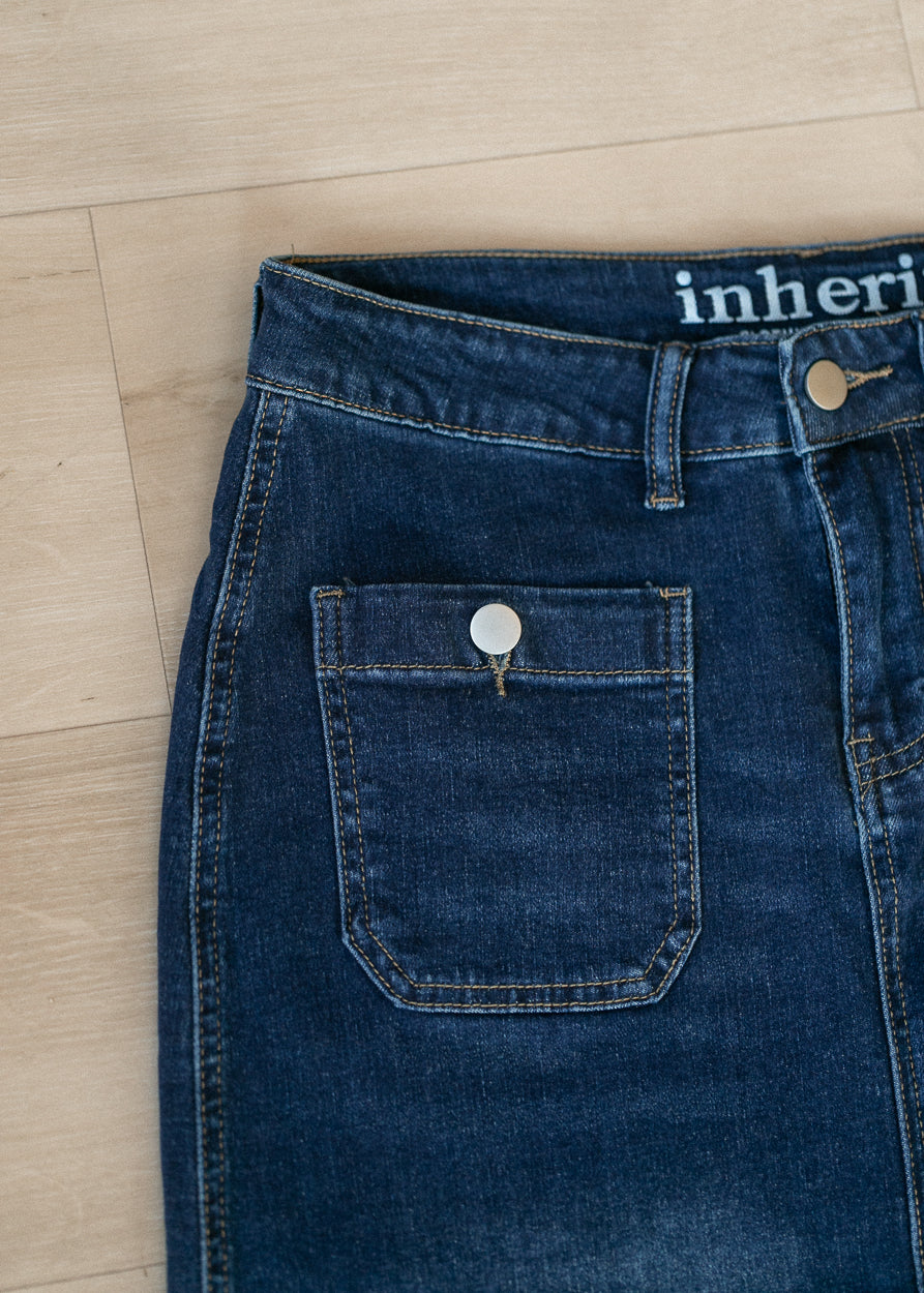 Close-up of dark blue denim jeans laid flat on a light wood floor, showing the waistband, belt loops, front pocket, and a partial brand wordmark reading 'inheri'.