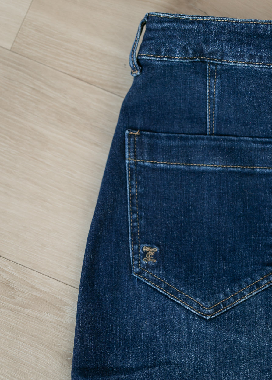 Close-up of the back pocket of dark blue denim jeans laid flat on a light wood floor, showing a small embroidered logo on the pocket.