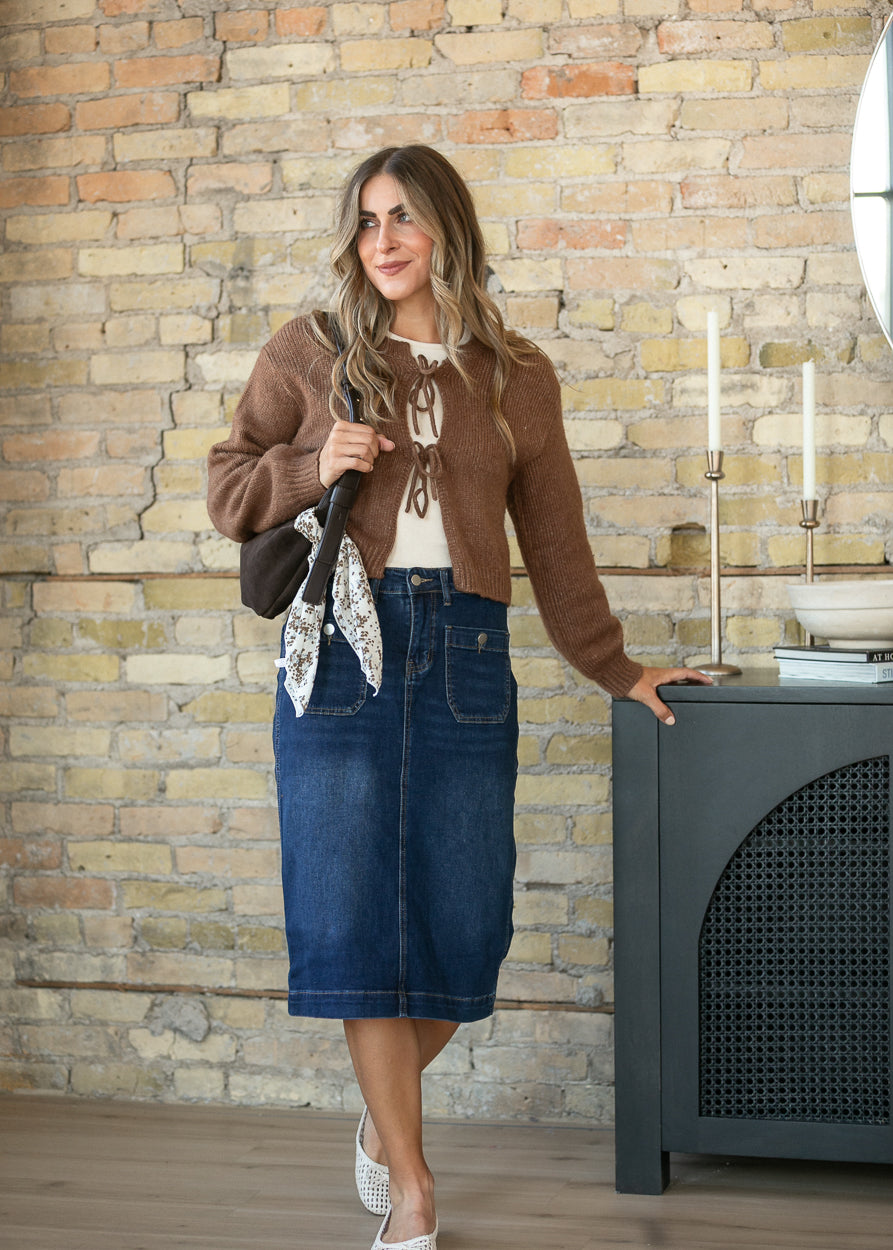 A woman stands in a room with a brick wall, wearing a brown knit cardigan over a white top, a blue denim midi skirt, and white wedge sandals. She is holding a patterned scarf and standing next to a dark gray cabinet.