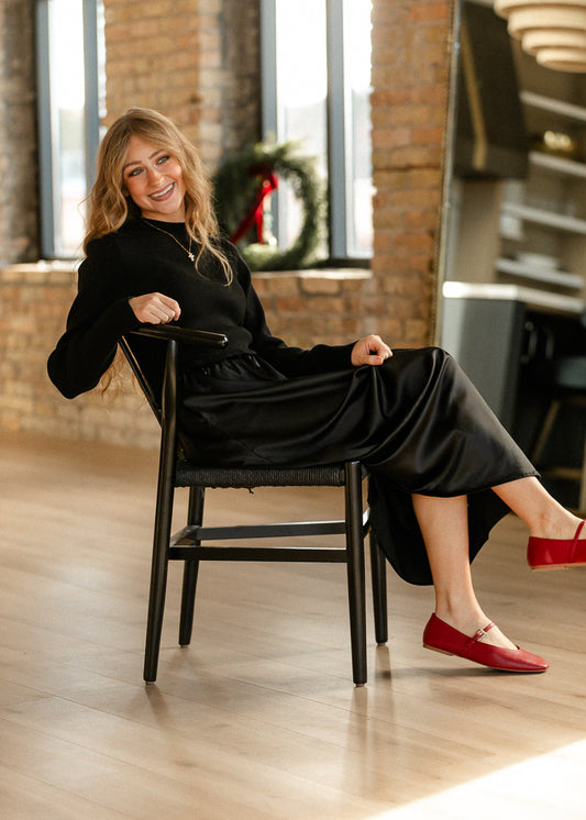 Ember black maxi dress worn seated, showing its long sleeve sweater bodice and flowing skirt with red Mary Jane flats.